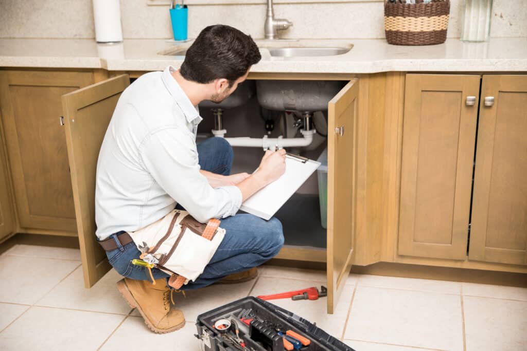 Rear view of a male plumber writing a repair order while crouching in front of a kitchen sink